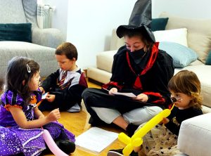 En un salón se encuentra una bruja sentada con las piernas cruzadas mientras lee a los niños que la rodean. Hay 3 niños, dos de ellos disfrazados (Harry Potter y Bruja), quienes escuchan atentamente a la bruja quien le ha dado globos.
